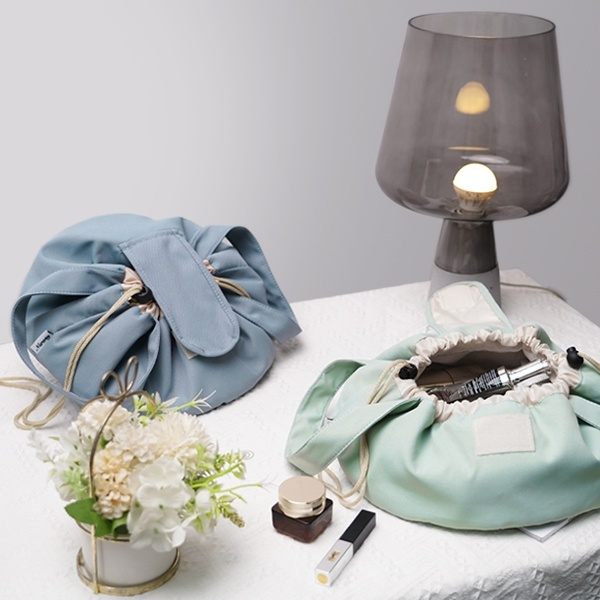 Light blue drawstring bag, green fabric bag with makeup items, small plant, and gray lamp on a white surface.