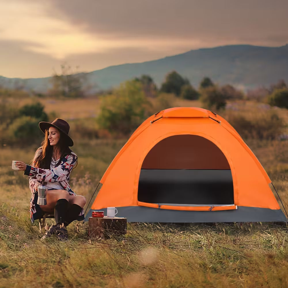 Woman sitting next to an orange tent in a field with mountains in the background