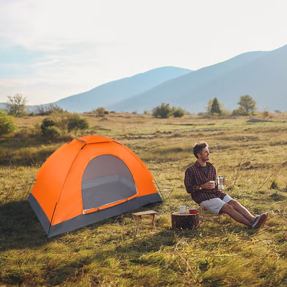 Man sitting by an orange tent in a grassy field with mountains in the background