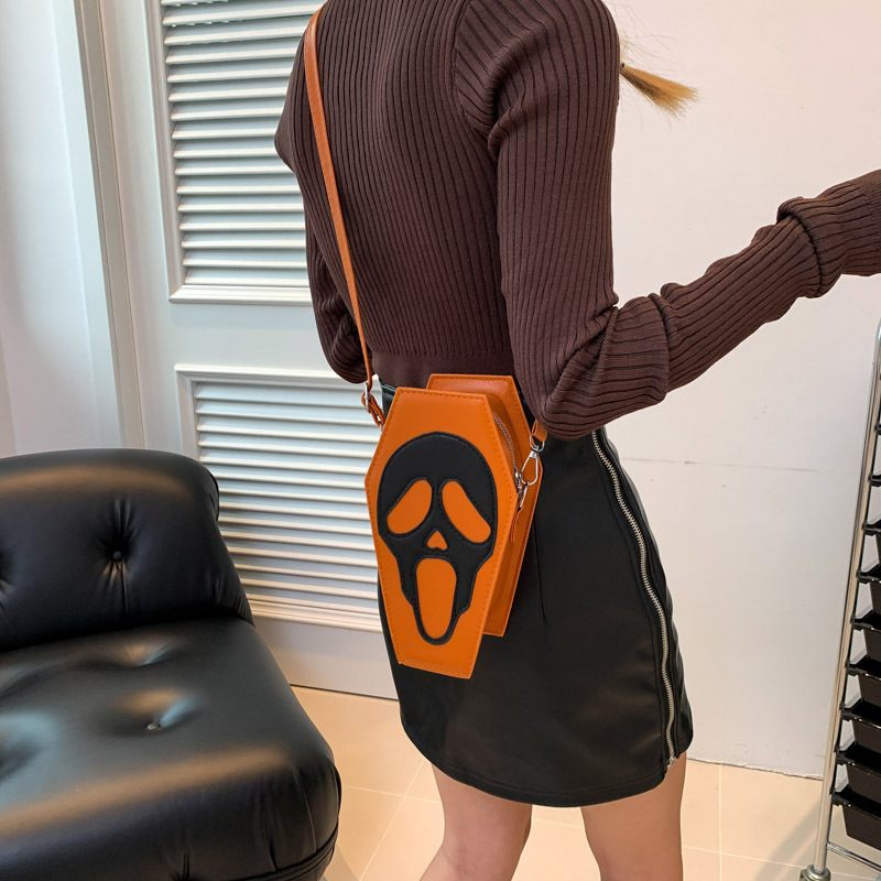 Person wearing a brown ribbed sweater and black skirt with an orange coffin-shaped bag featuring a ghost face design.
