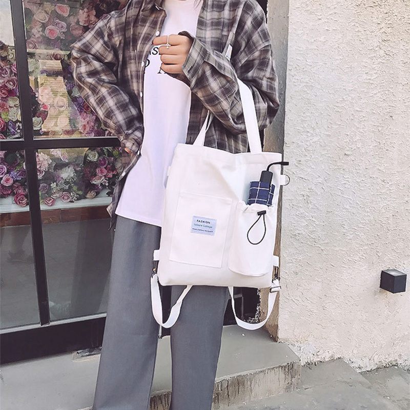 Person holding a white tote bag with a visible brand label, standing against a textured wall.