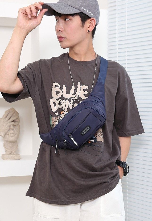 Person wearing a brown t-shirt with a graphic design, holding a navy blue waist bag, and wearing a gray cap indoors.