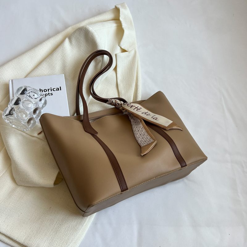 Brown handbag with brown handle and strap on a white surface with a book and decorative item.
