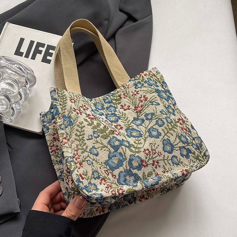 Floral patterned bag held by a hand on a white surface with a gray jacket and glass object in the background.