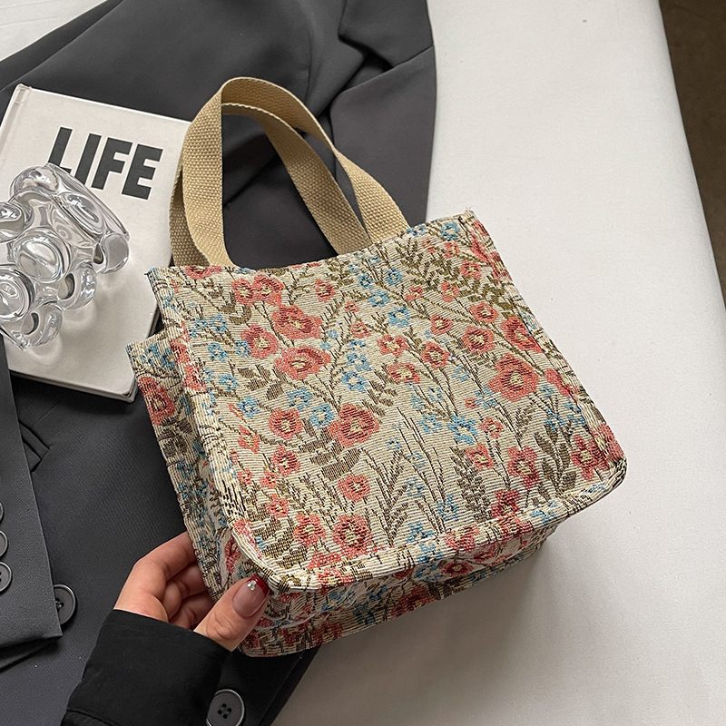 Floral patterned handbag held by a hand on a white surface with a magazine and glass in the background.