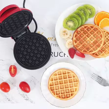 Waffle maker with a cooked waffle on a plate, surrounded by fruits on a white surface.