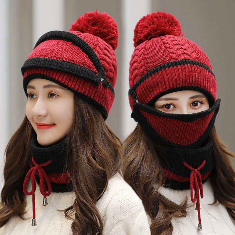 Red and black knitted hat with pom-pom and face mask worn by a person against a neutral background
