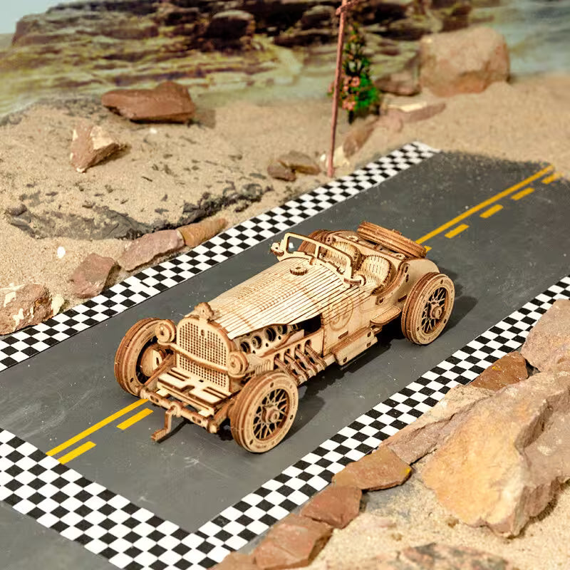 Wooden model car on a miniature road with checkered pattern and rocky landscape.