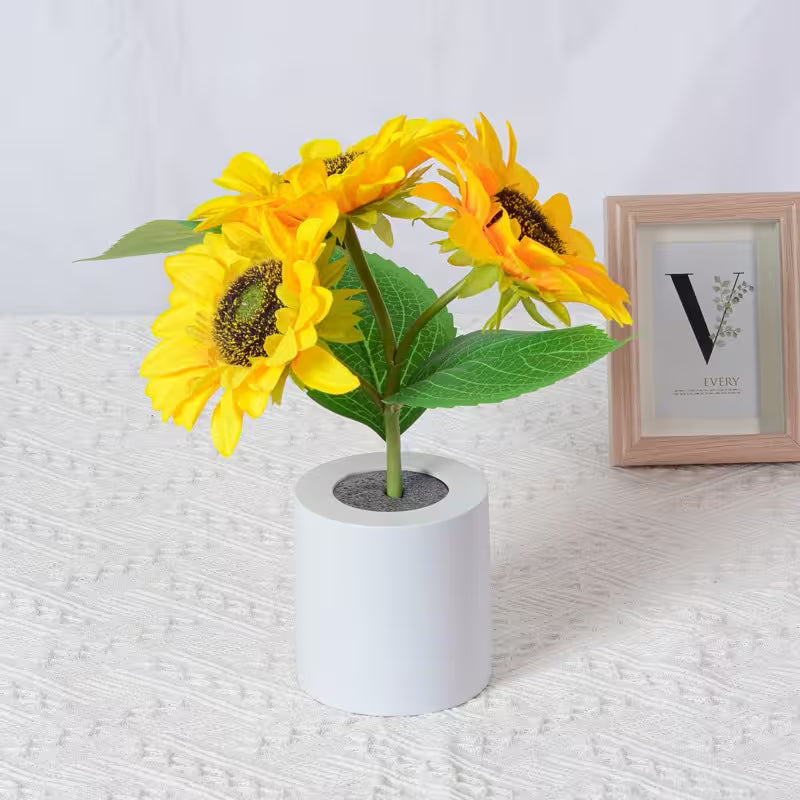 Artificial sunflower plant in a white pot on a textured surface with a framed picture in the background.