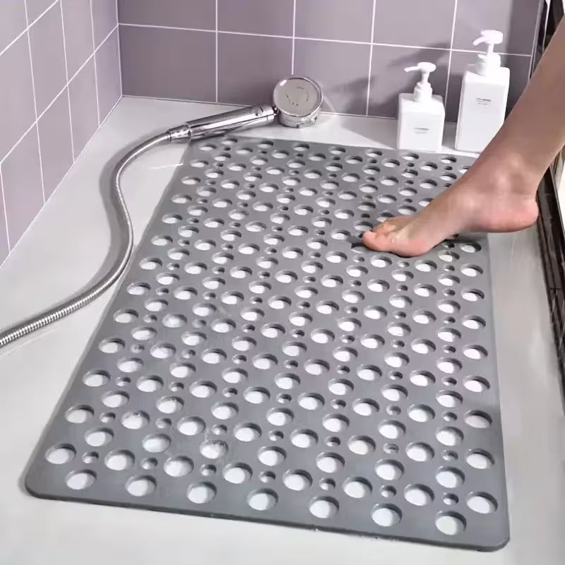 Gray perforated bath mat on a bathroom floor with a shower head and bottles in the background.