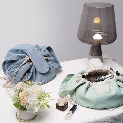 Light blue drawstring bag, green fabric bag with makeup items, small plant, and gray lamp on a white surface.