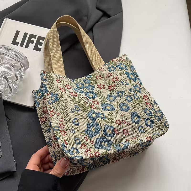 Floral patterned bag held by a hand on a white surface with a magazine and glass in the background.