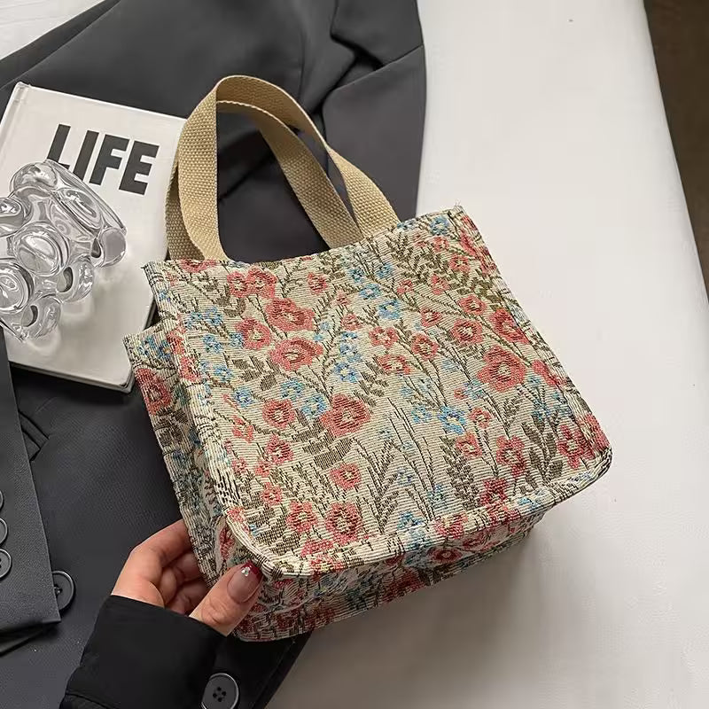 Floral patterned handbag held by a hand on a desk with a magazine and glassware in the background.