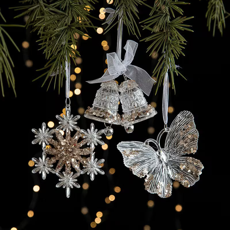 Decorative Christmas ornaments including a butterfly, snowflake, and bell hanging on a tree with blurred lights in the background.