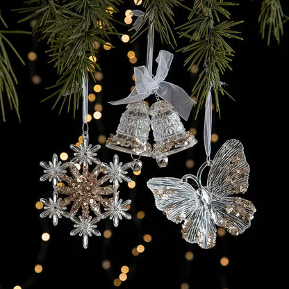 Decorative Christmas ornaments including a butterfly, snowflake, and bell hanging on a tree with blurred lights in the background.