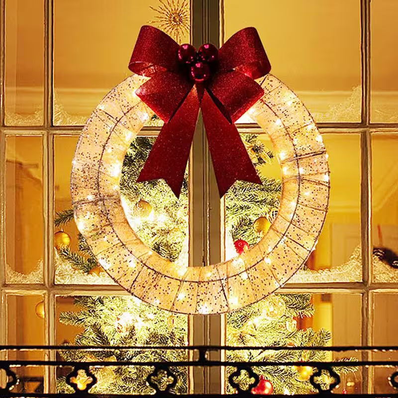 Decorative wreath with lights and a red bow in front of a window with Christmas tree reflections.
