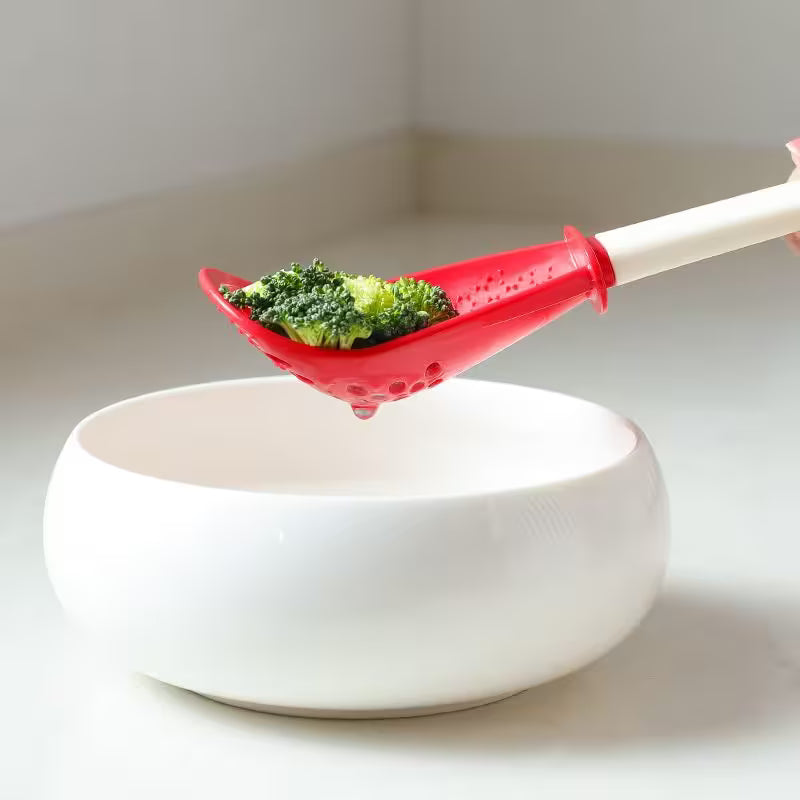 Red silicone spoon with green vegetables over a white bowl on a light background