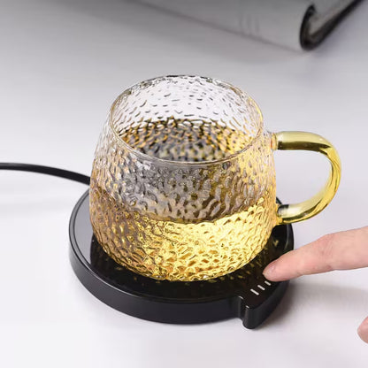 Glass mug with gold interior on a black heating pad, being held by a hand.