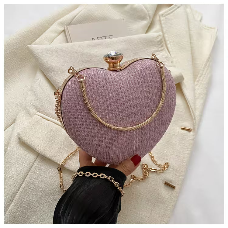 Heart-shaped pink clutch with gold chain held by a hand against a white background