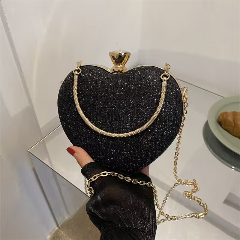 Black glittery heart-shaped handbag with gold chain held by a hand on a reflective surface.