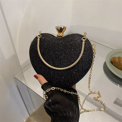 Black glittery heart-shaped handbag with gold chain held by a hand on a reflective surface.