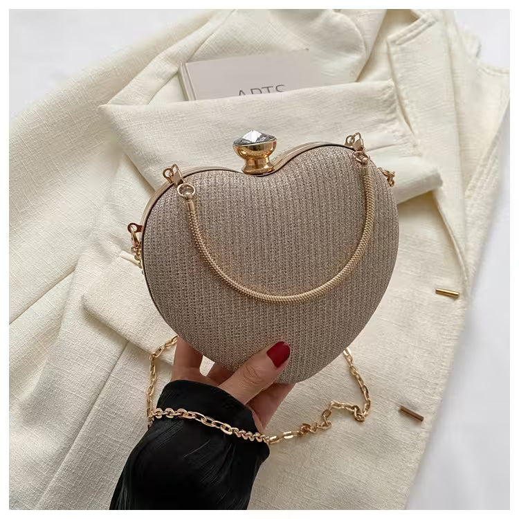 Heart-shaped clutch bag with gold chain held by a hand against a white coat background