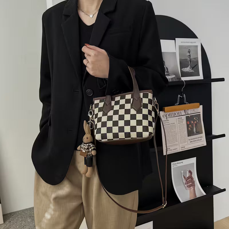 Person holding a checkered handbag with a small plush toy, standing in front of a black shelving unit with magazines.