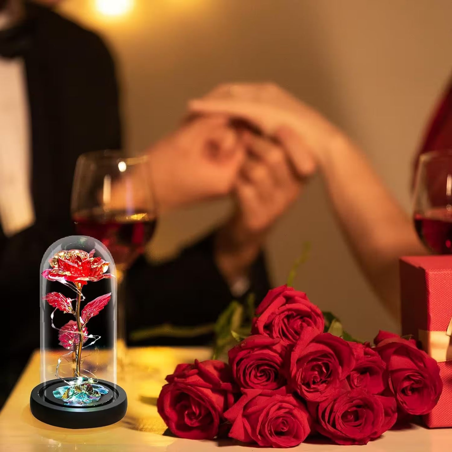 Bouquet of red roses and a glass dome with a rose on a table with people in the background holding hands.