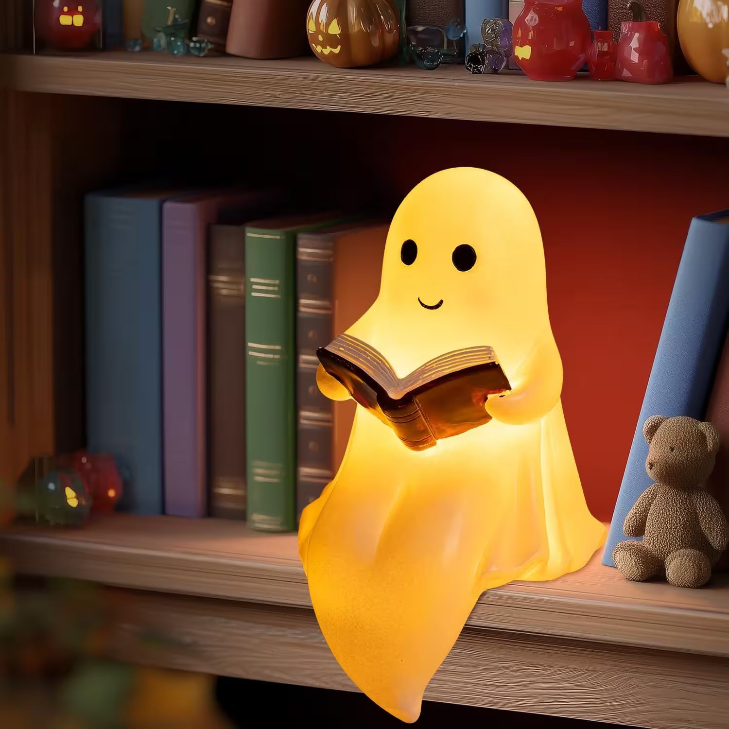 Ghost-shaped night light reading a book on a shelf with books and a teddy bear.