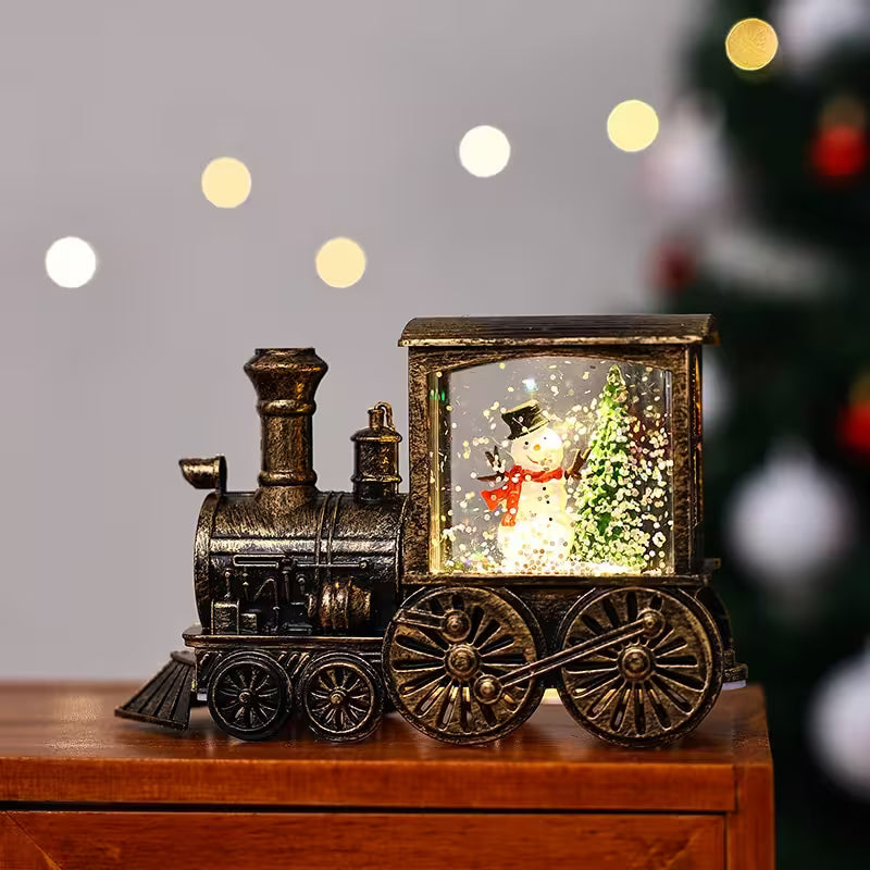 Decorative train-shaped water lamp with a snowman inside, on a wooden surface with blurred lights in the background.