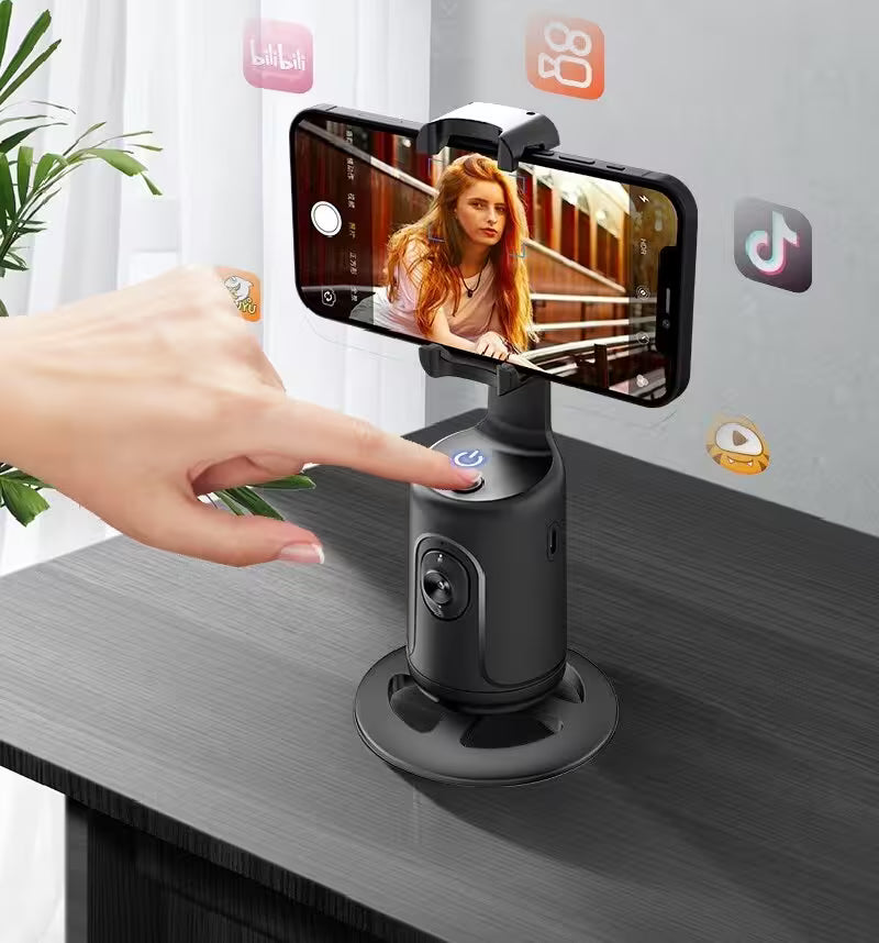 Hand using a smartphone with a gimbal attachment on a desk, with social media app icons in the background.