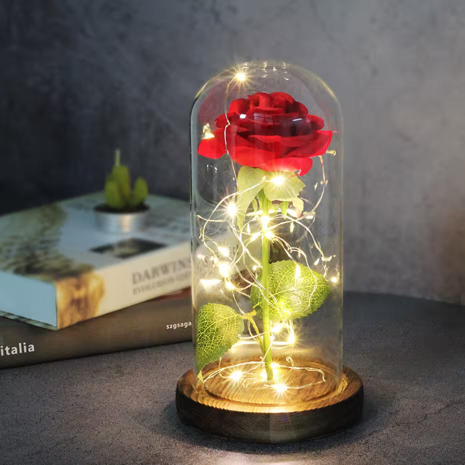 Red rose in a glass dome with fairy lights on a wooden base, placed on a surface with a blurred book in the background.