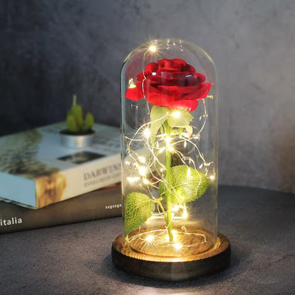 Red rose in a glass dome with fairy lights on a wooden base, placed on a surface with a blurred book in the background.