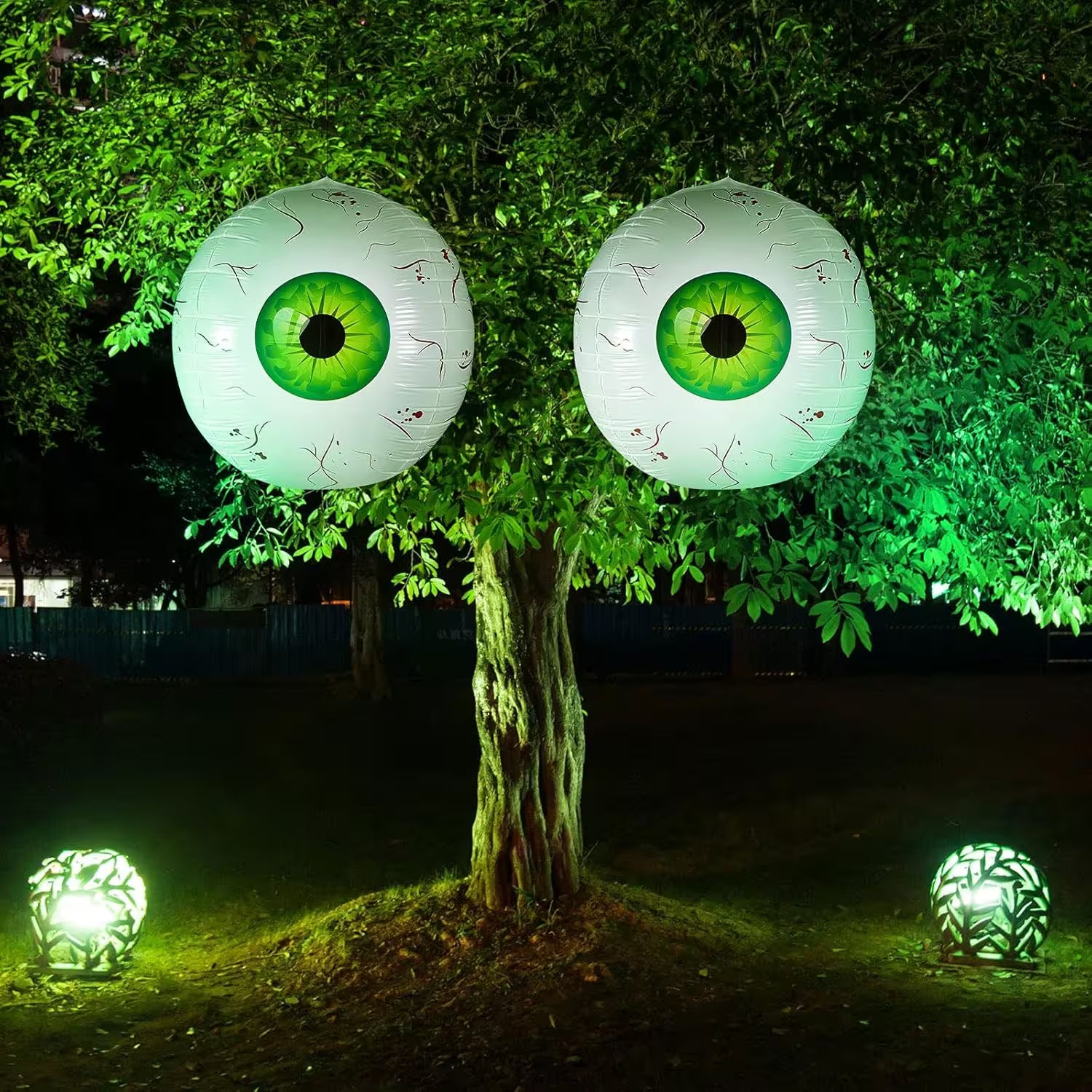 Two large eye-shaped lanterns hanging from a tree with additional lanterns on the ground at night.