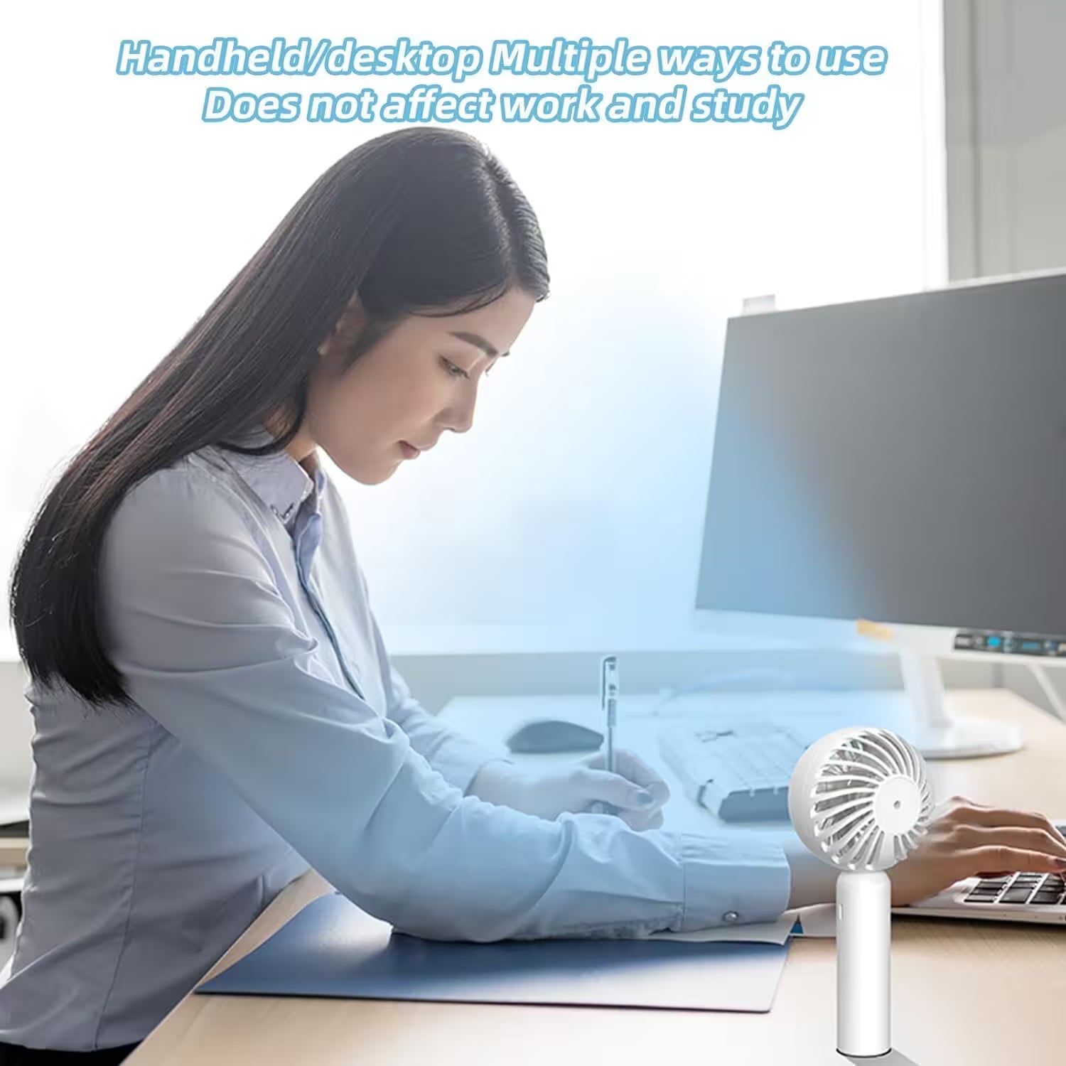 Woman working at a desk with a handheld fan in front of her computer