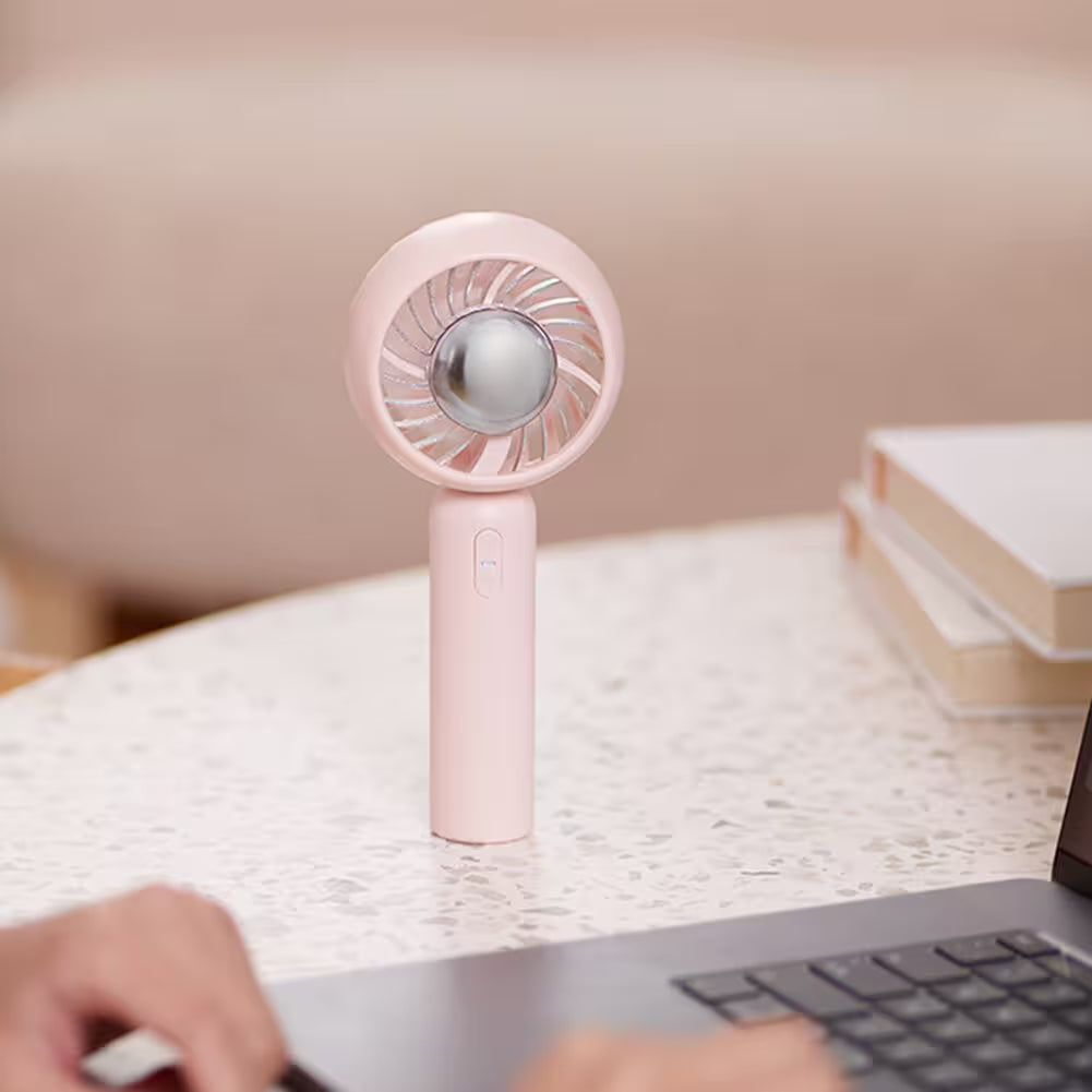 Pink handheld fan on a desk with a laptop and books in the background