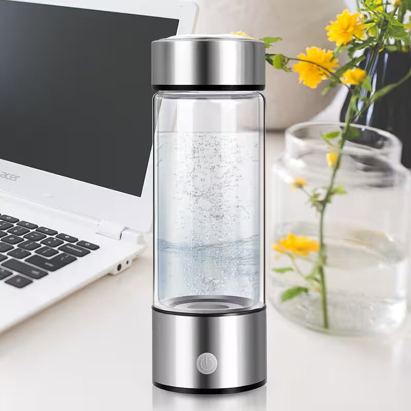 Clear glass water bottle with silver top and base on a desk with laptop and flowers
