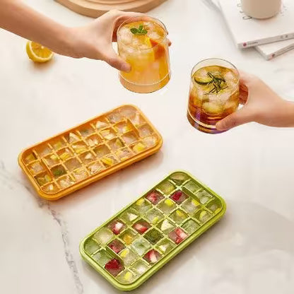 Two ice cube trays with colorful ice cubes on a marble surface, accompanied by glasses of iced drinks.