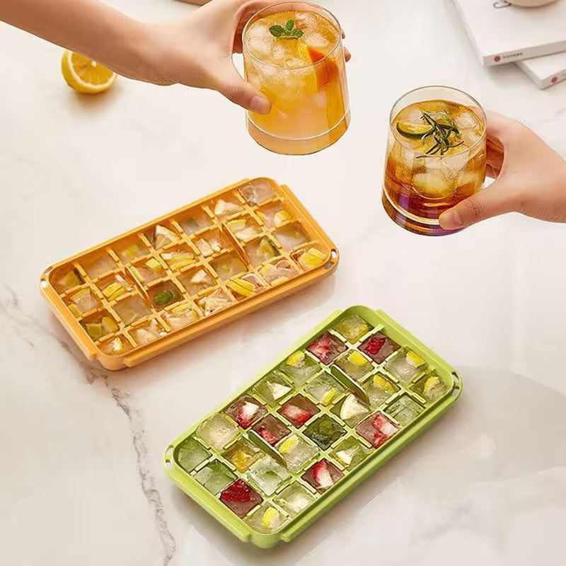 Two ice cube trays with colorful ice cubes, one yellow and one green, on a marble surface with drinks being held.
