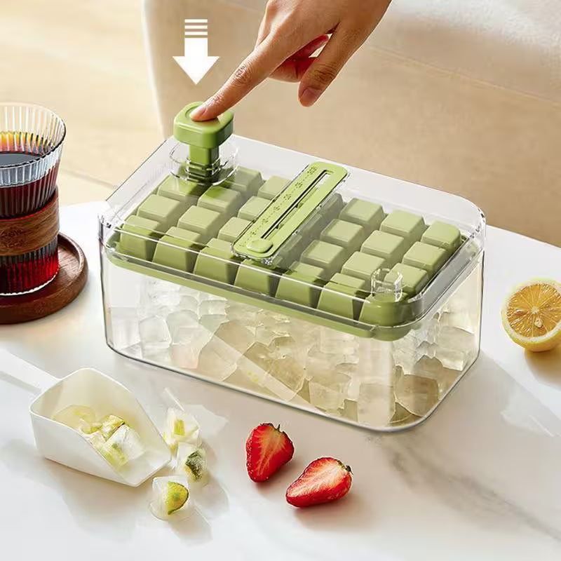 Green ice cube tray with clear lid on a kitchen counter, surrounded by fruits and a glass of juice.