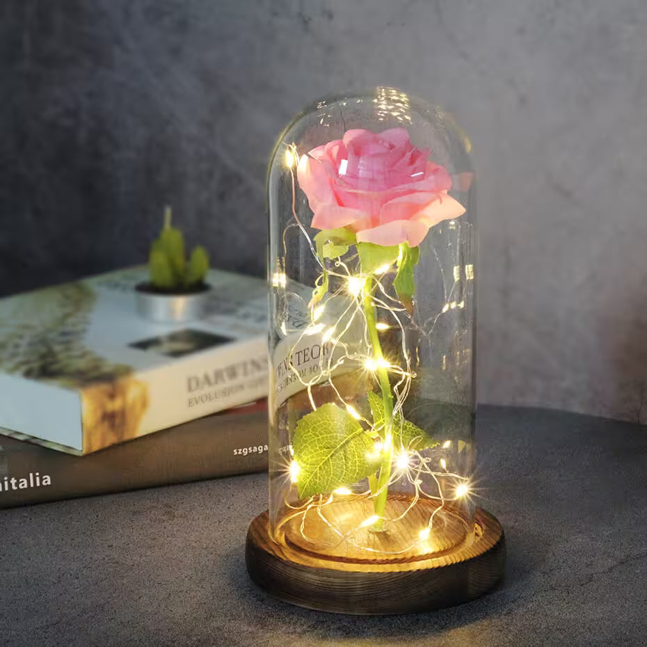 Pink rose in a glass dome with LED lights on a wooden base, placed on a surface with a blurred book in the background.