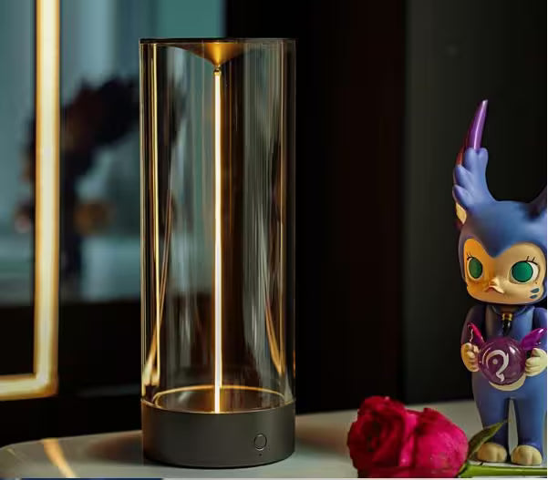 Decorative glass cylinder with a glowing interior on a surface next to a colorful figurine and a rose.