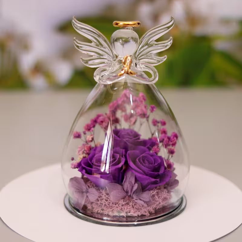 Glass dome with an angel figurine containing purple flowers on a white surface.