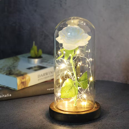White rose in a glass dome with LED lights on a wooden base, placed on a surface with a book in the background.