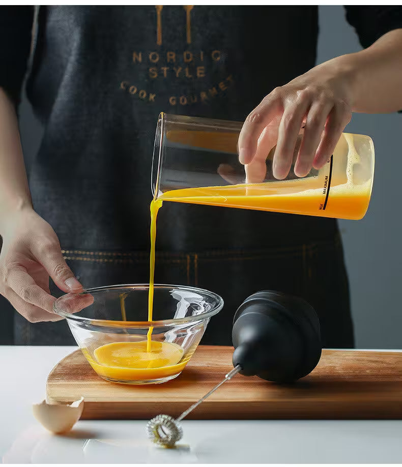 Person pouring a yellow liquid from a measuring container into a glass bowl on a wooden cutting board.