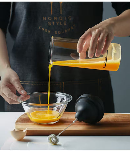 Person pouring a yellow liquid from a measuring container into a glass bowl on a wooden cutting board.
