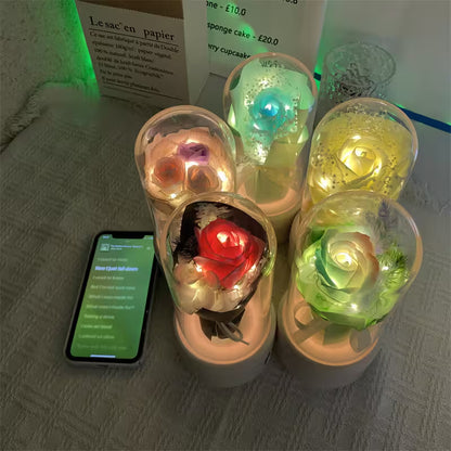 Colorful LED-lit roses in transparent cups on a textured surface with a phone displaying text.