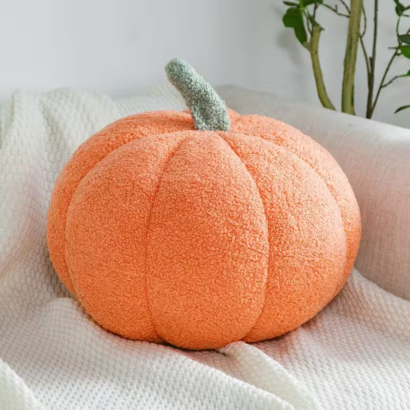 Plush pumpkin pillow on a white blanket with a plant in the background