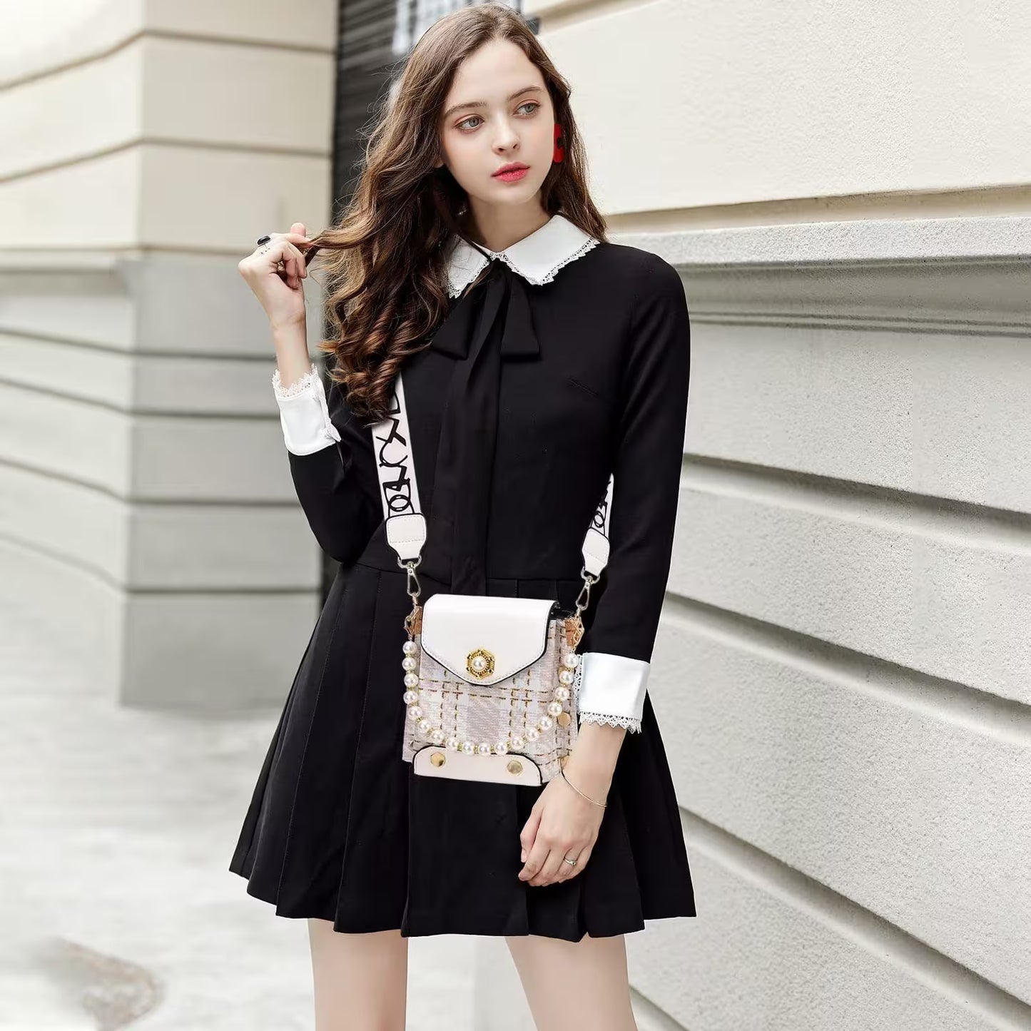 Woman wearing a black dress with a white collar and holding a decorative handbag against a light-colored wall.
