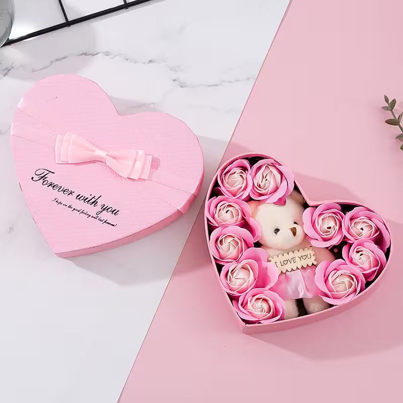 Heart-shaped pink gift box with a teddy bear and flowers on a white marble surface.